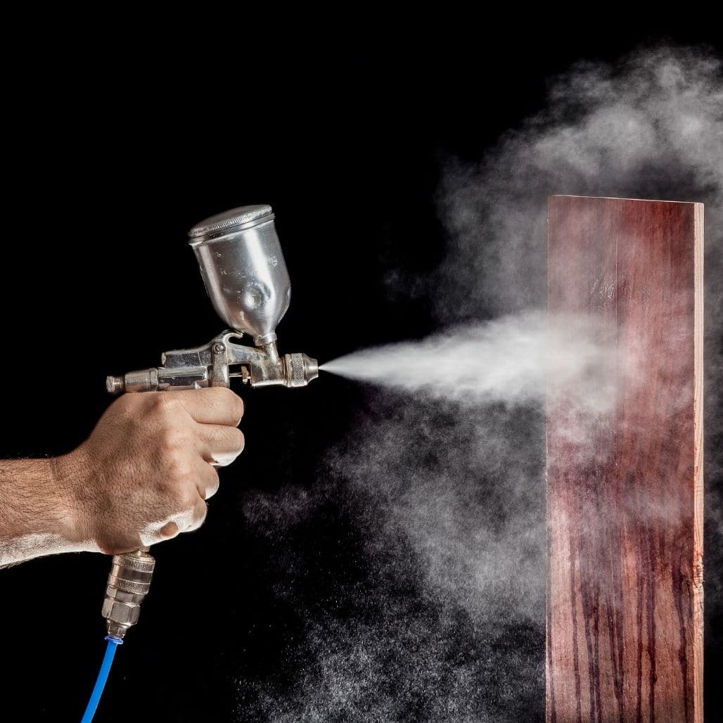 Worker holding a paint spray gun discharging atomised mist onto a wooden panel — isocyanate exposure during spraying