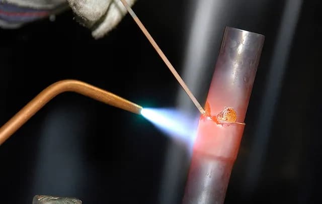 Brazing torch joining a copper pipe with filler rod and visible flame — metal fume and lead exposure source
