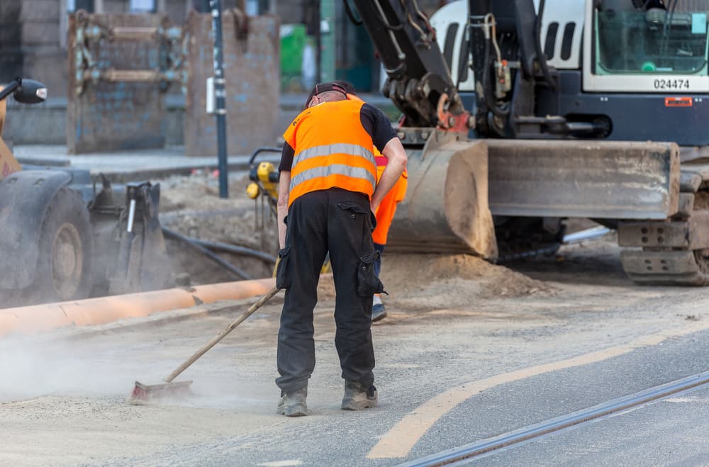 High-vis worker sweeping cement dust on a street-works site beside an excavator — respirable crystalline silica exposure